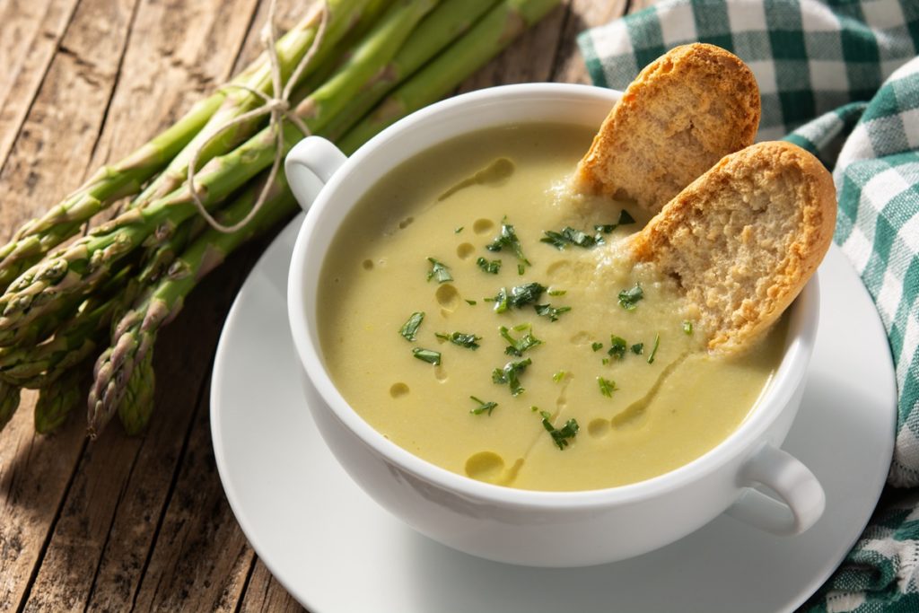 Supă cremă de sparanghel în bol alb, cu două felii de pâine, alături de o legătură de sparanghel