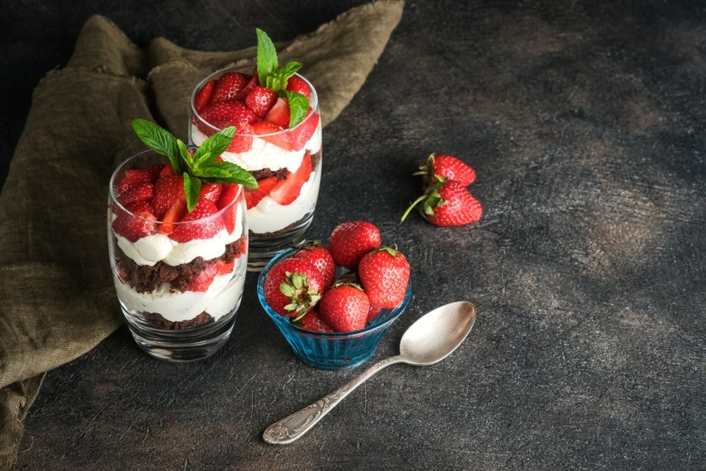 Două porții de Trifles cu căpșuni și biscuiți cu ciocolată în pahare, alături de o linguriță și un bol cu căpșuni