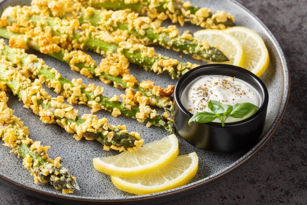 Sparanghel în crustă de porumb pe o farfurie cu felii de lămâie și un bol cu sos de iaurt