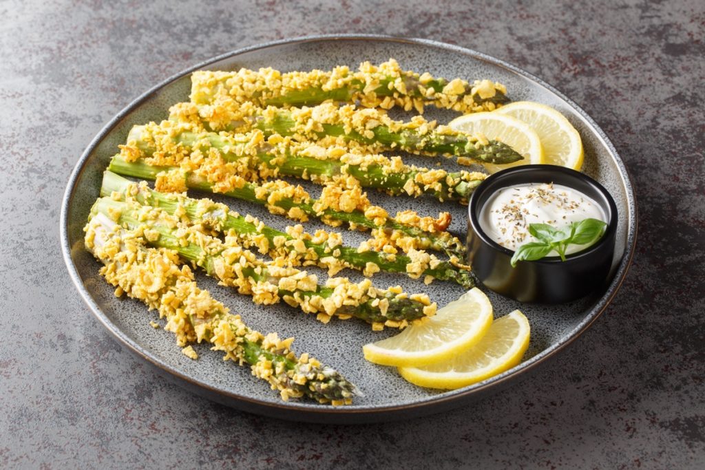 Sparanghel în crustă de porumb pe o farfurie cu felii de lămâie și un bol cu sos de iaurt