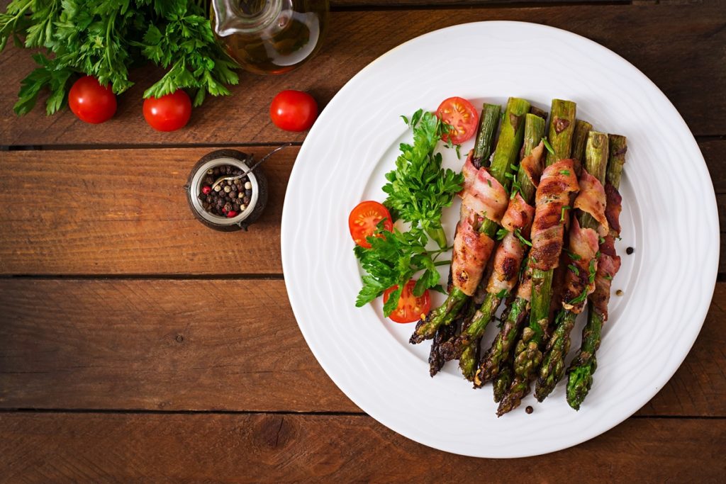 Sparanghel înfășurat în bacon pe o farfurie cu roșii cherry și verdeață