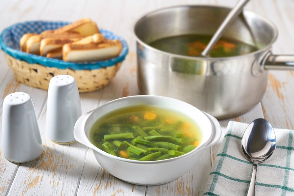 Supă de fasole verde cu morcovi în bol și în cratiță cu polonic, alături de un coș cu felii de pâine