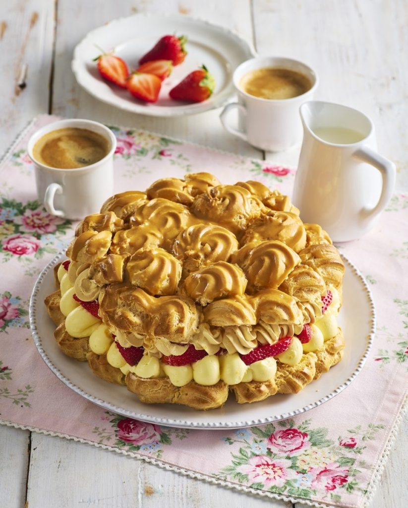 Tort Ecler cu cremă de banane, căpșuni și caramel, alături de două cești cu cafea, una cu lapte și un bol cu căpșuni