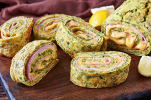 Ruladă din omletă cu dovlecei, umplută cu șuncă și cașcaval, porționată pe un tocător de lemn