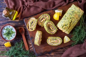 Ruladă din omletă cu dovlecei, umplută cu șuncă și cașcaval, porționată pe un tocător de lemn