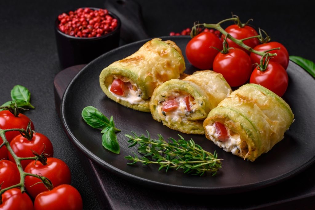 Trei rulouri de dovlecei cu brânză și roșii pe platou, alături de roșii proaspete și crenguțe de rozmarin
