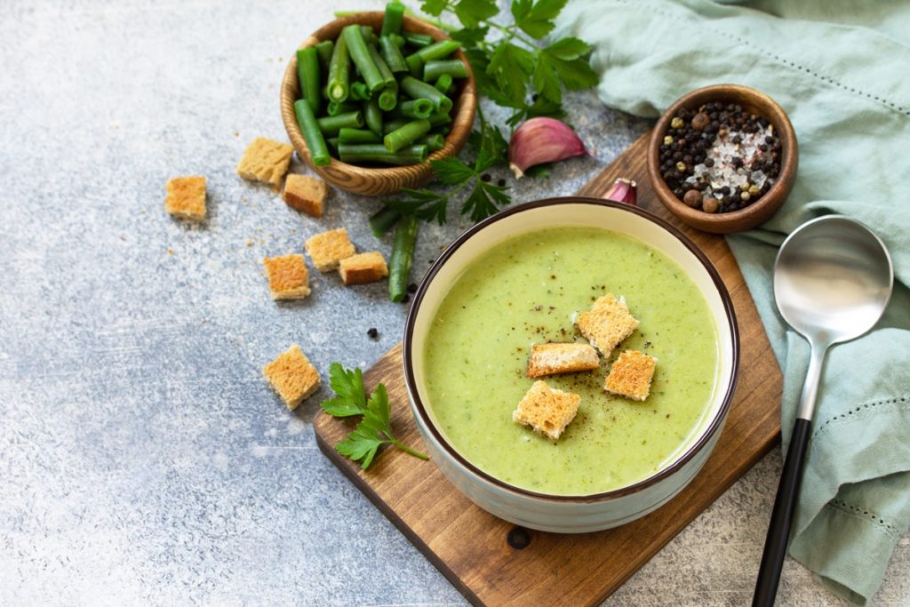 Supă cremă de fasole verde și crutoane în bol, alături de un bol cu păstăi verzi și unul cu condimente