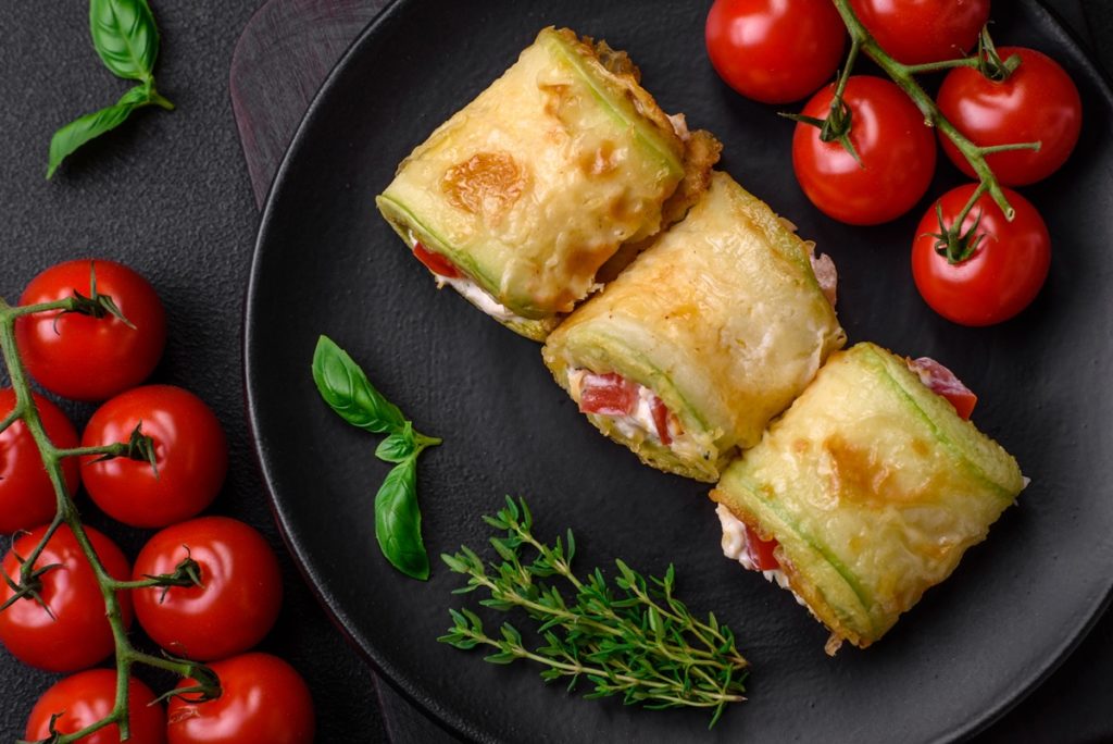 Trei rulouri de dovlecei cu brânză și roșii pe platou, alături de roșii proaspete și crenguțe de rozmarin