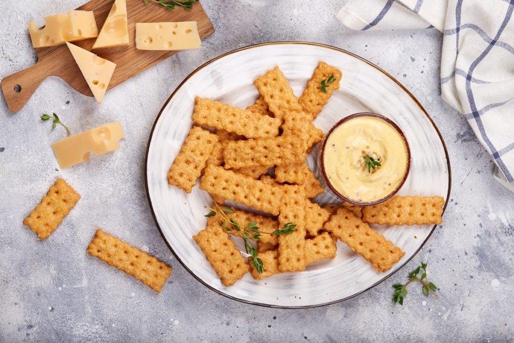 Biscuiți crocanți cu cașcaval afumat pe o farfurie, alături de un bol cu cremă de brânză