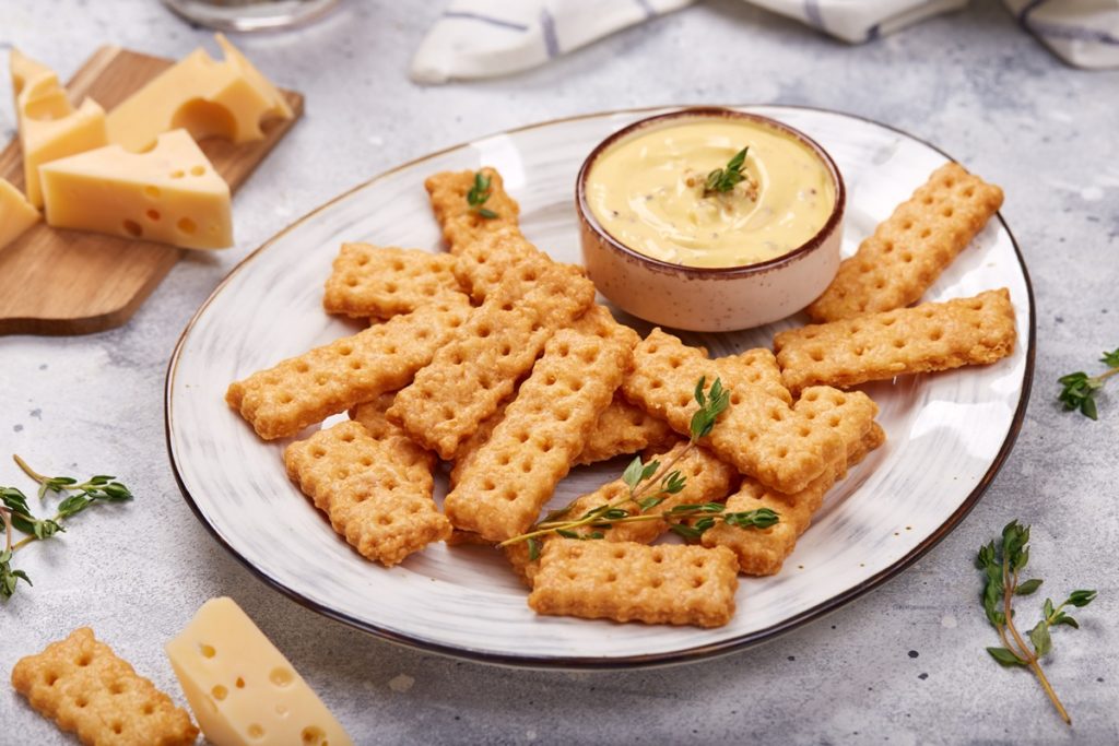 Biscuiți crocanți cu cașcaval afumat pe o farfurie, alături de un bol cu cremă de brânză