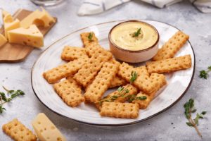 Biscuiți crocanți cu cașcaval afumat pe o farfurie, alături de un bol cu cremă de brânză