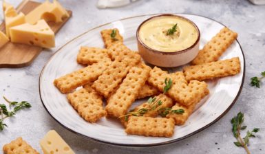 Biscuiți crocanți cu cașcaval afumat pe o farfurie, alături de un bol cu cremă de brânză