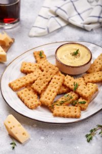 Biscuiți crocanți cu cașcaval afumat pe o farfurie, alături de un bol cu cremă de brânză