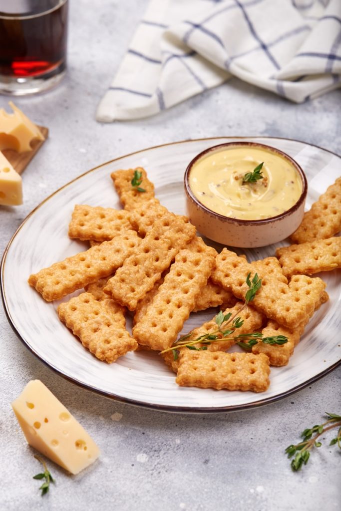 Biscuiți crocanți cu cașcaval afumat pe o farfurie, alături de un bol cu cremă de brânză
