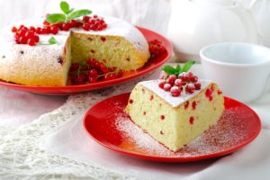 Porție de pandișpan pufos cu coacăze roșii pe o farfurie, alături de platoul cu pandișpan secționat