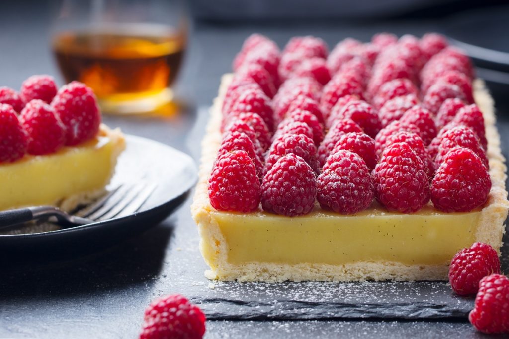 Tartă cu lămâie și zmeură pe un platou și o porție pe farfurie, alături de un pahar cu băutură