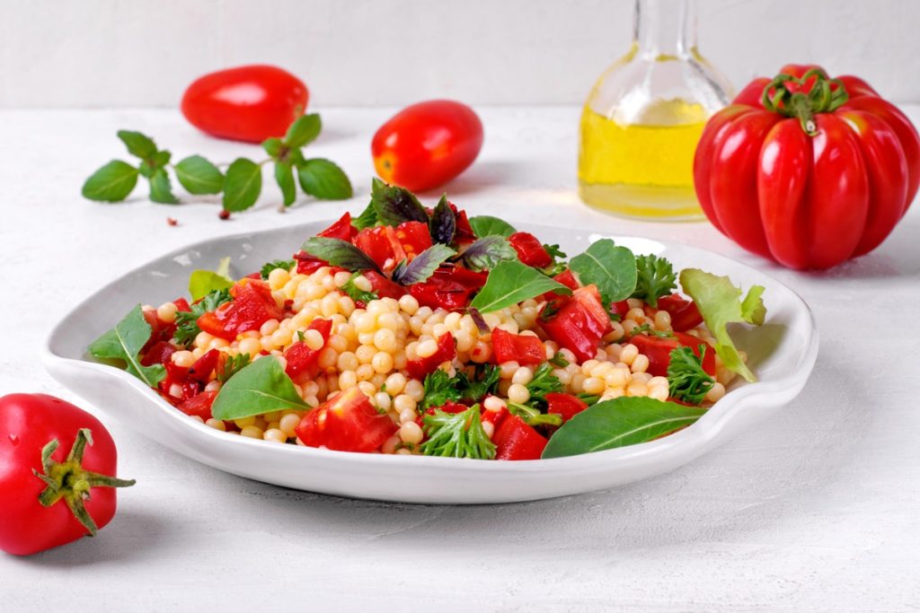 Salată de bulgur cu roșii și ardei în bol, alături de roșii, o sticlă cu ulei și frunze verzi