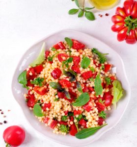 Salată de bulgur cu roșii și ardei în bol, alături de roșii, o sticlă cu ulei și boabe de piper