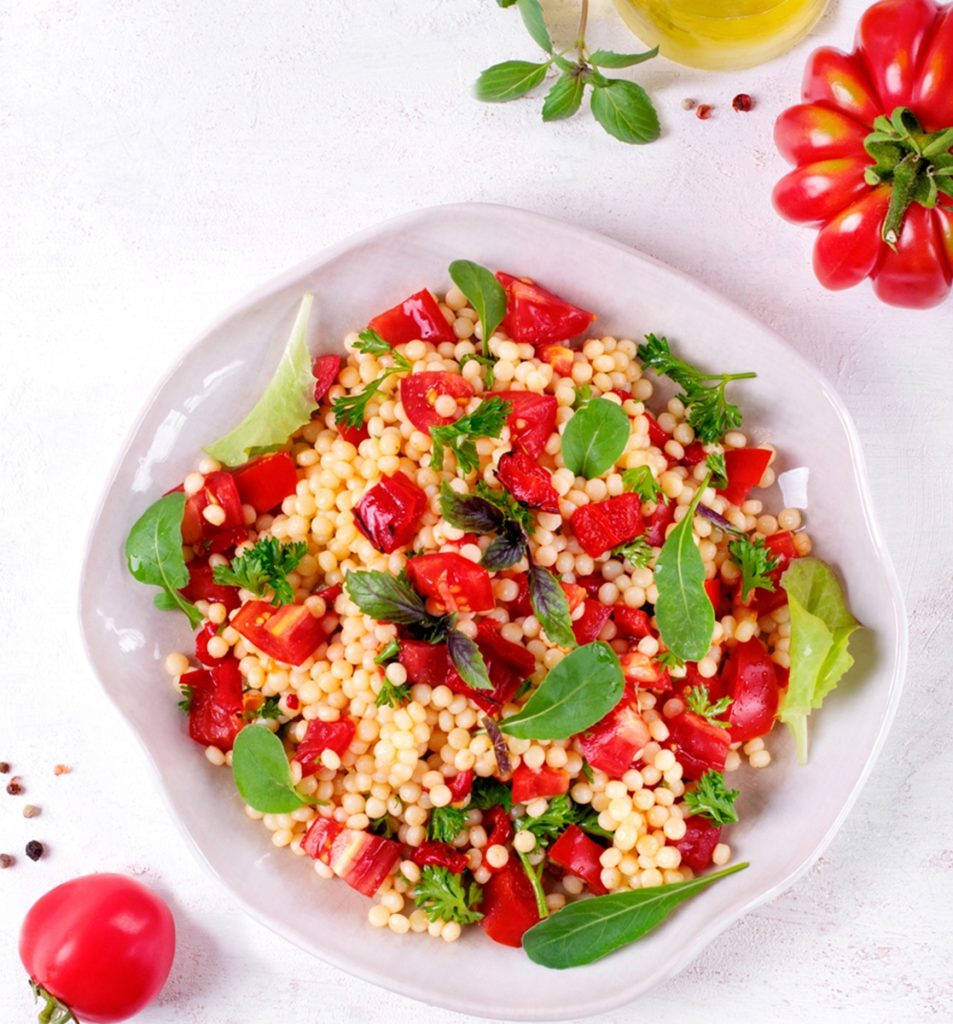 Salată de bulgur cu roșii și ardei în bol, alături de roșii, o sticlă cu ulei și boabe de piper