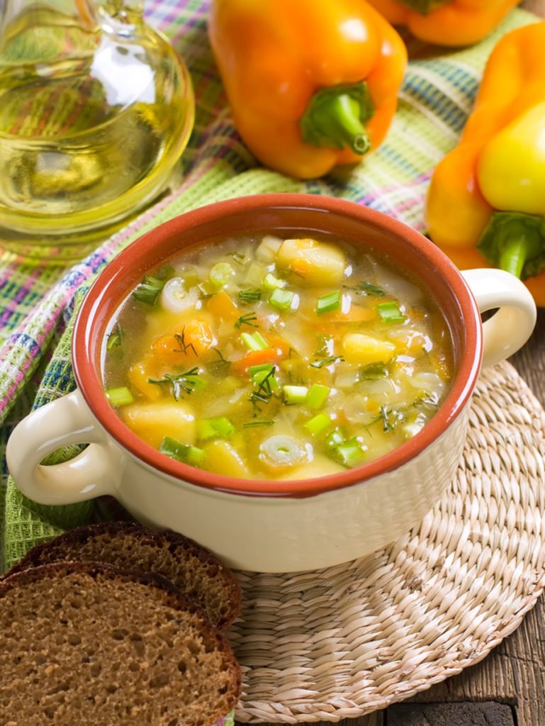 Supă deasă din legume de vară în bol, alături de ardei, o sticlă de ulei și felii de pâine