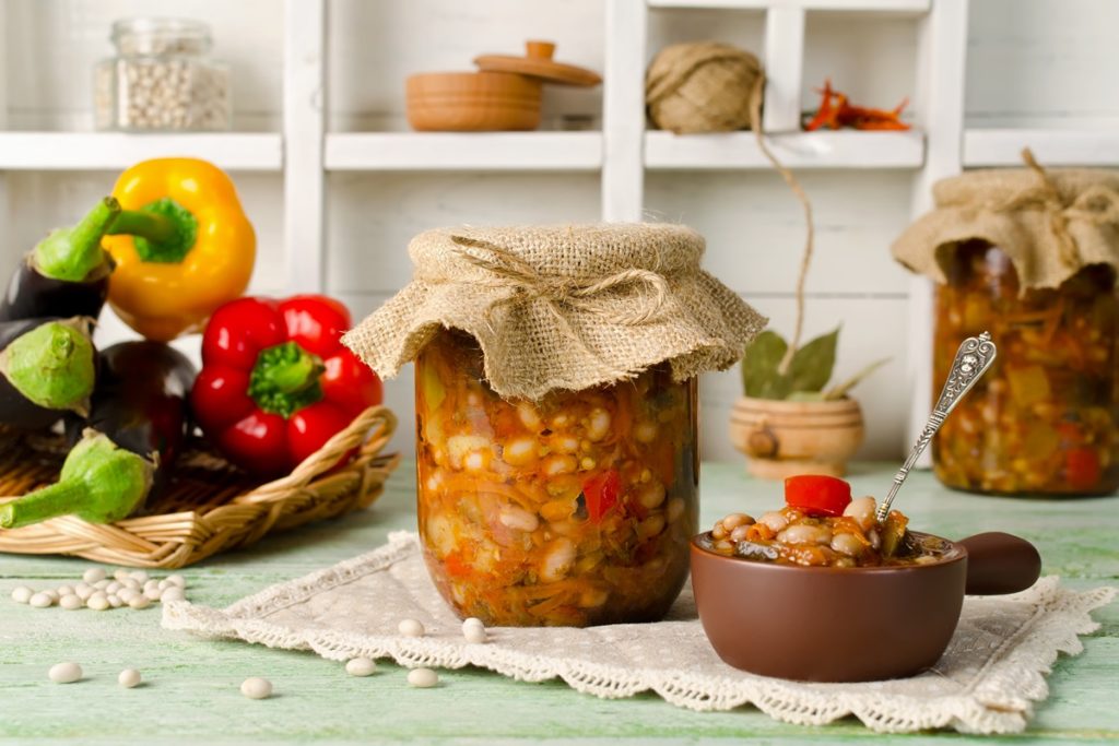 Două borcane și un bol cu zacuscă de fasole, alături de un bol cu vinete și ardei