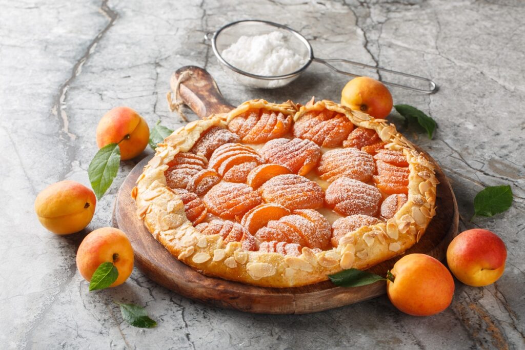 Galette cu caise în formă de evantai pe un tocător de lemn, alături de caise proaspete și un bol cu zahăr