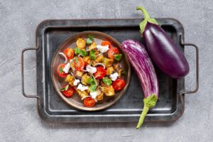 Salată de vinete prăjite, cu roșii și brânză într-o farfurie pe o tavă, alături de două vinete