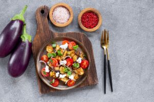 Salată de vinete prăjite, cu roșii și brânză într-o farfurie pe un tocător, alături de furculiță și cuțit, un bol cu sare și unul cu piper roșu
