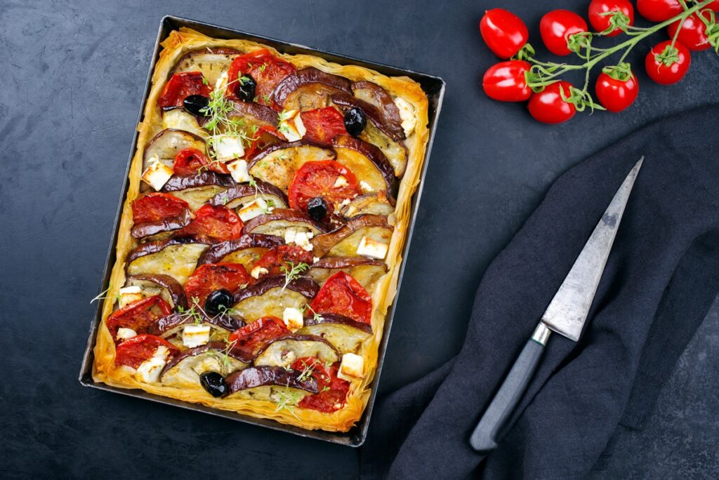 Tartă cu vinete, roșii și feta, în foi de plăcintă, în tava de copt, alături de un cuțit și crenguțe cu roșii