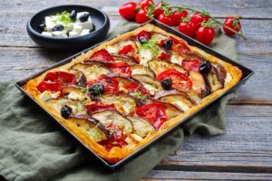 Tartă cu vinete, roșii și feta, în foi de plăcintă, în tava de copt, alături de un bol cu brânză și măsline și crenguțe cu roșii