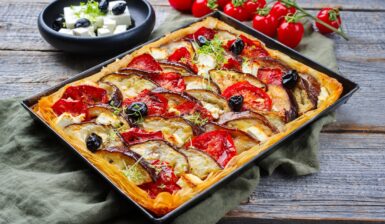 Tartă cu vinete, roșii și feta, în foi de plăcintă, în tava de copt, alături de un bol cu brânză și măsline și crenguțe cu roșii