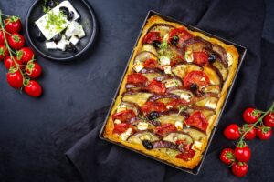 Tartă cu vinete, roșii și feta, în foi de plăcintă, în tava de copt, alături de un bol cu brânză și măsline și crenguțe cu roșii