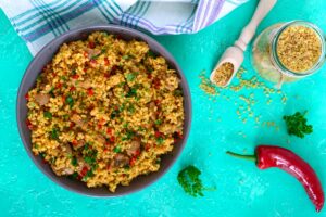 Bulgur cu carne de pui și legume în bol, alături de un bol și o lingură cu bulgur și un ardei roșu
