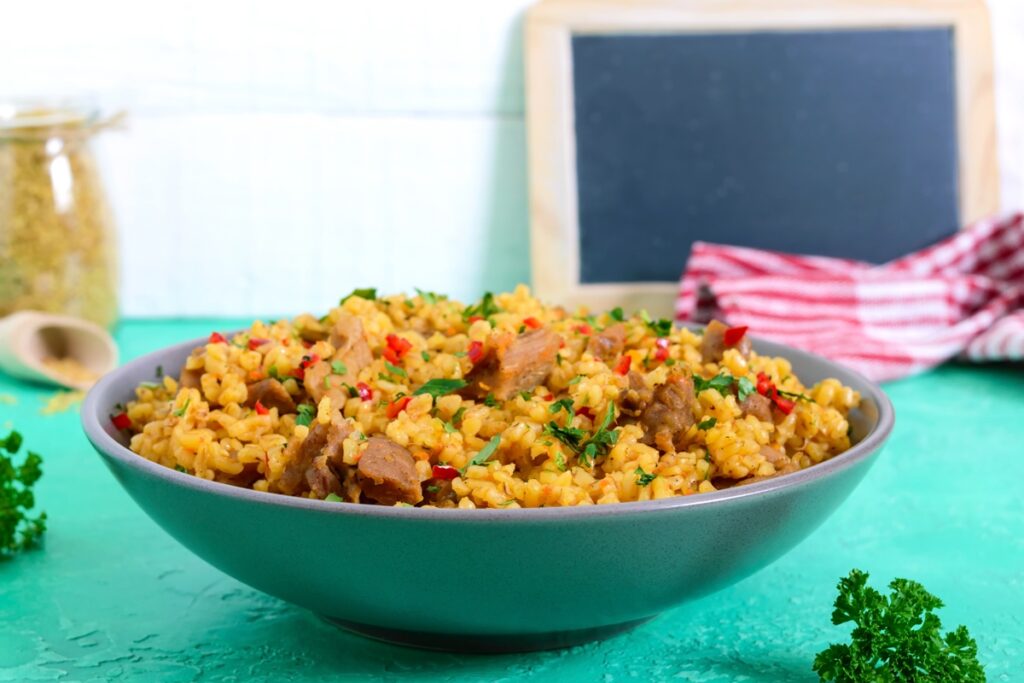 Bulgur cu carne de pui și legume în bol, un borcan cu bulgur, o lingură de lemn și frunze de pătrunjel