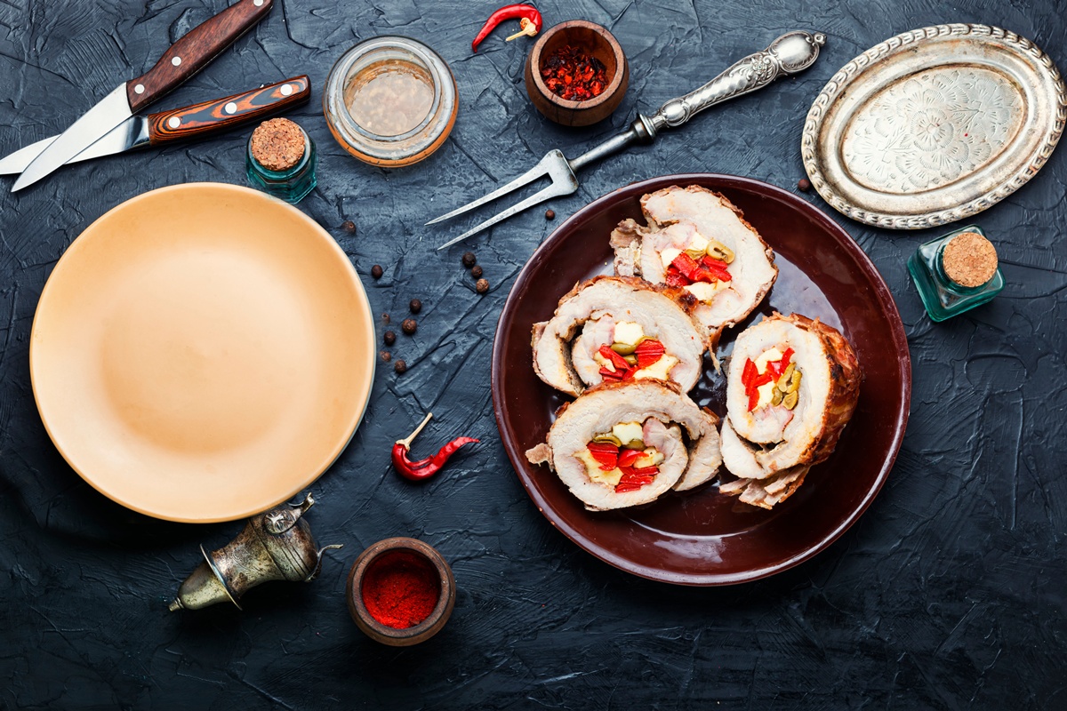 Patru porții de Ruladă de porc cu gogoșari murați, măsline verzi și cașcaval pe un platou, tacâmuri, condimente, o farfurie și un platou