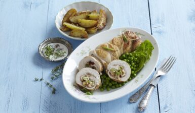 Ruladă de curcan cu brânză cremoasă, mazăre și roșii uscate porționată pe un platou cu mazăre, alături de un bol cu cartofi și unul cu sos