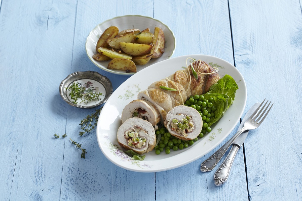 Ruladă de curcan cu brânză cremoasă, mazăre și roșii uscate porționată pe un platou cu mazăre, alături de un bol cu cartofi și unul cu sos