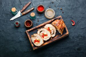 Ruladă de porc cu gogoșari murați, măsline verzi și cașcaval porționată pe un platou, alături de tacâmuri și condimente