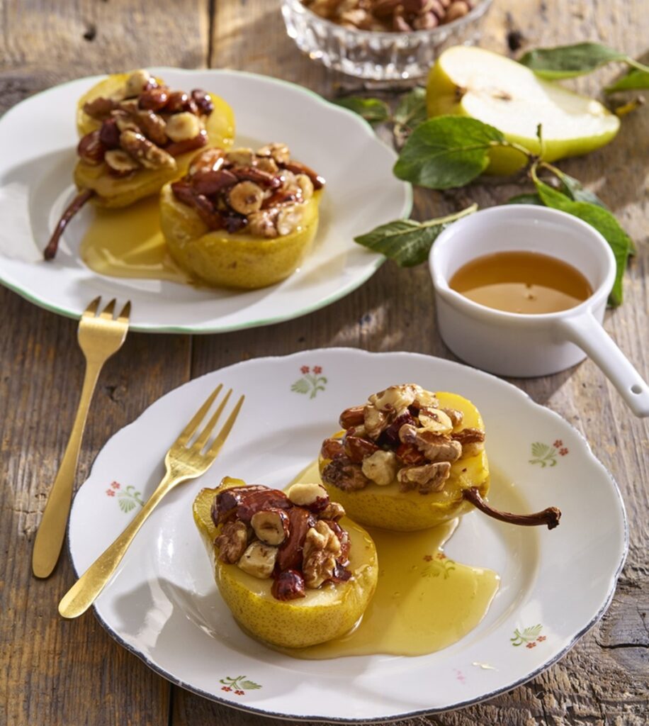 Jumătăți de pere coapte cu miere și umplute cu nuci și alune pe două farfurii cu furculițe și un bol cu miere