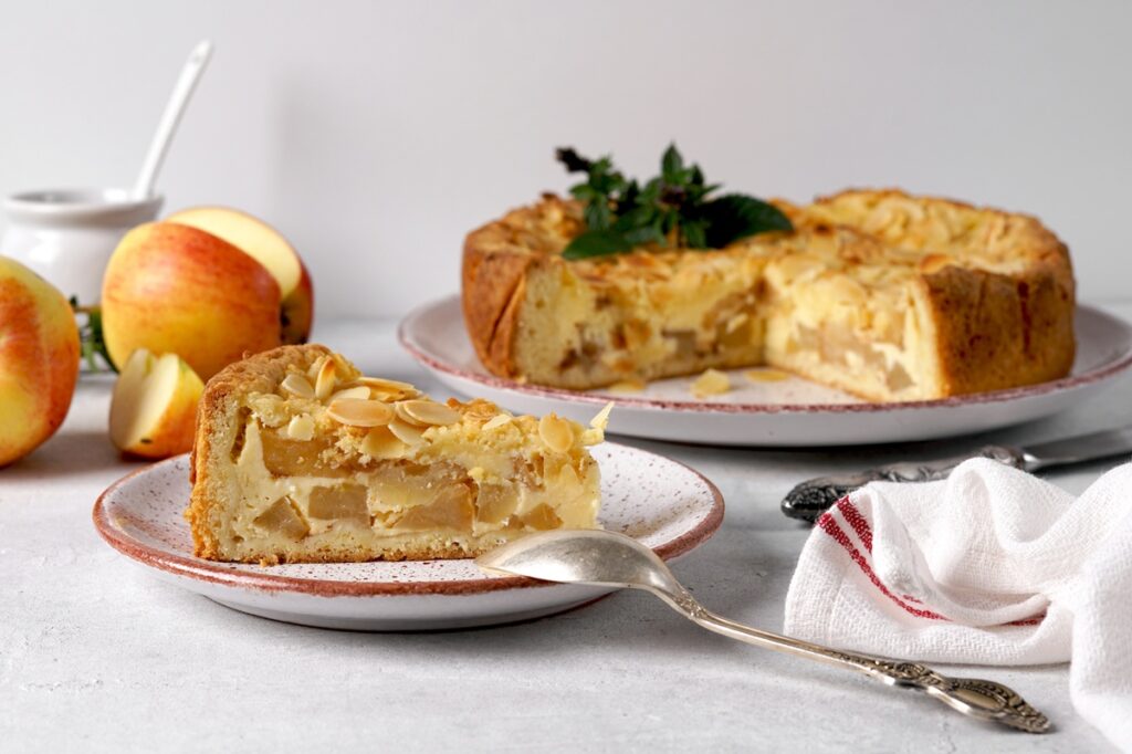 Prăjitură cu mere și vanilie, de post, porționată pe o farfurie, cu o linguriță, un cuțit, platoul cu prăjitura secționată și mere proaspete