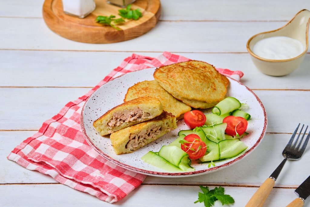 Plăcinte din aluat de cartofi cu carne tocată pe o farfurie, una secționată, servite cu salată de roșii și castraveți
