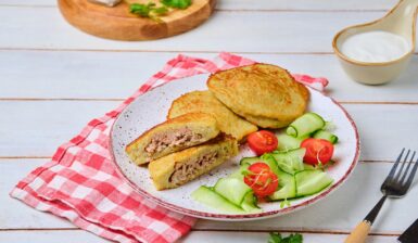 Plăcinte din aluat de cartofi cu carne tocată pe o farfurie, una secționată, servite cu salată de roșii și castraveți