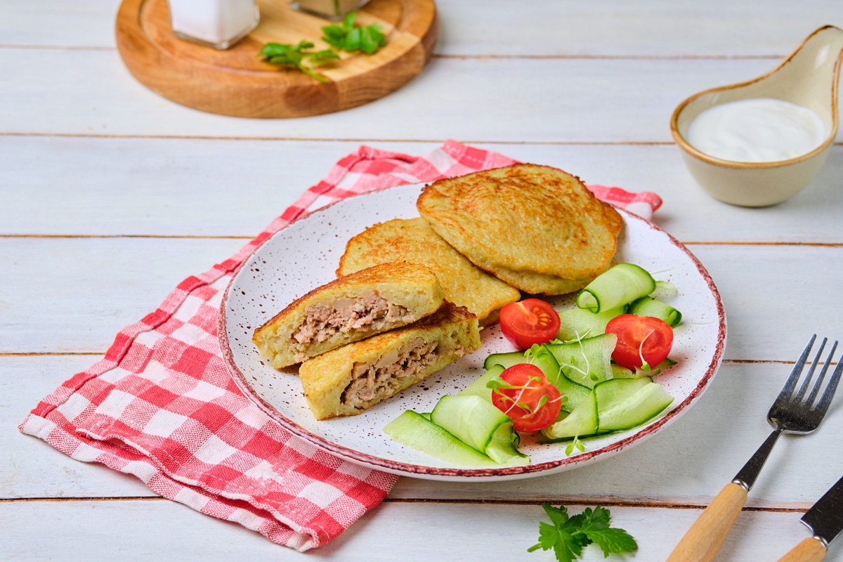 Plăcinte din aluat de cartofi cu carne tocată pe o farfurie, una secționată, servite cu salată de roșii și castraveți