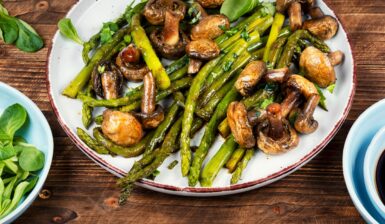 Sparanghel cu ciuperci la tigaie, în farfurie, alături de un bol cu frunze verzi