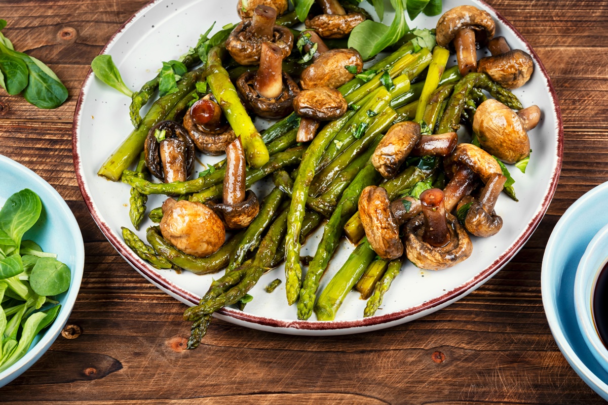 Sparanghel cu ciuperci la tigaie, în farfurie, alături de un bol cu frunze verzi