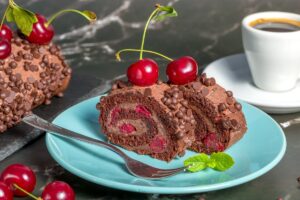 Porții de ruladă de cacao cu vișine și ciocolată pe farfurie, o ceașcă de cafea și rulada pe platou